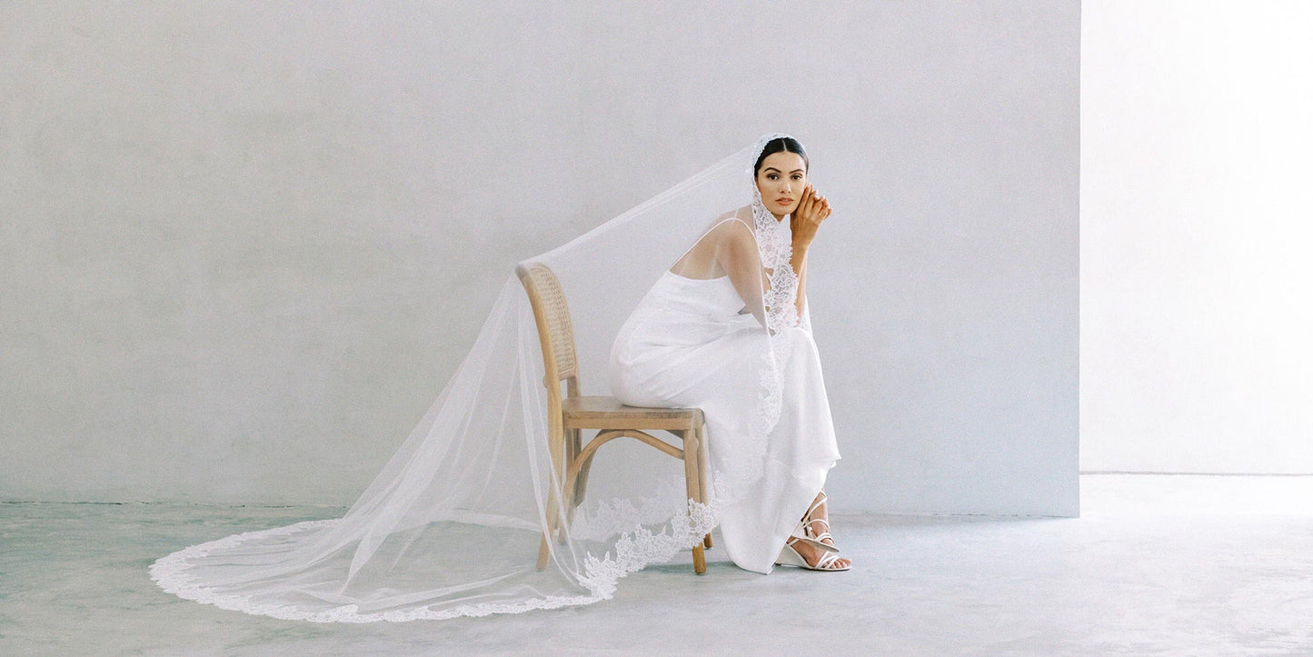 A model wearing a plain modern wedding dress and a long lace mantilla veil sitting on a chair 