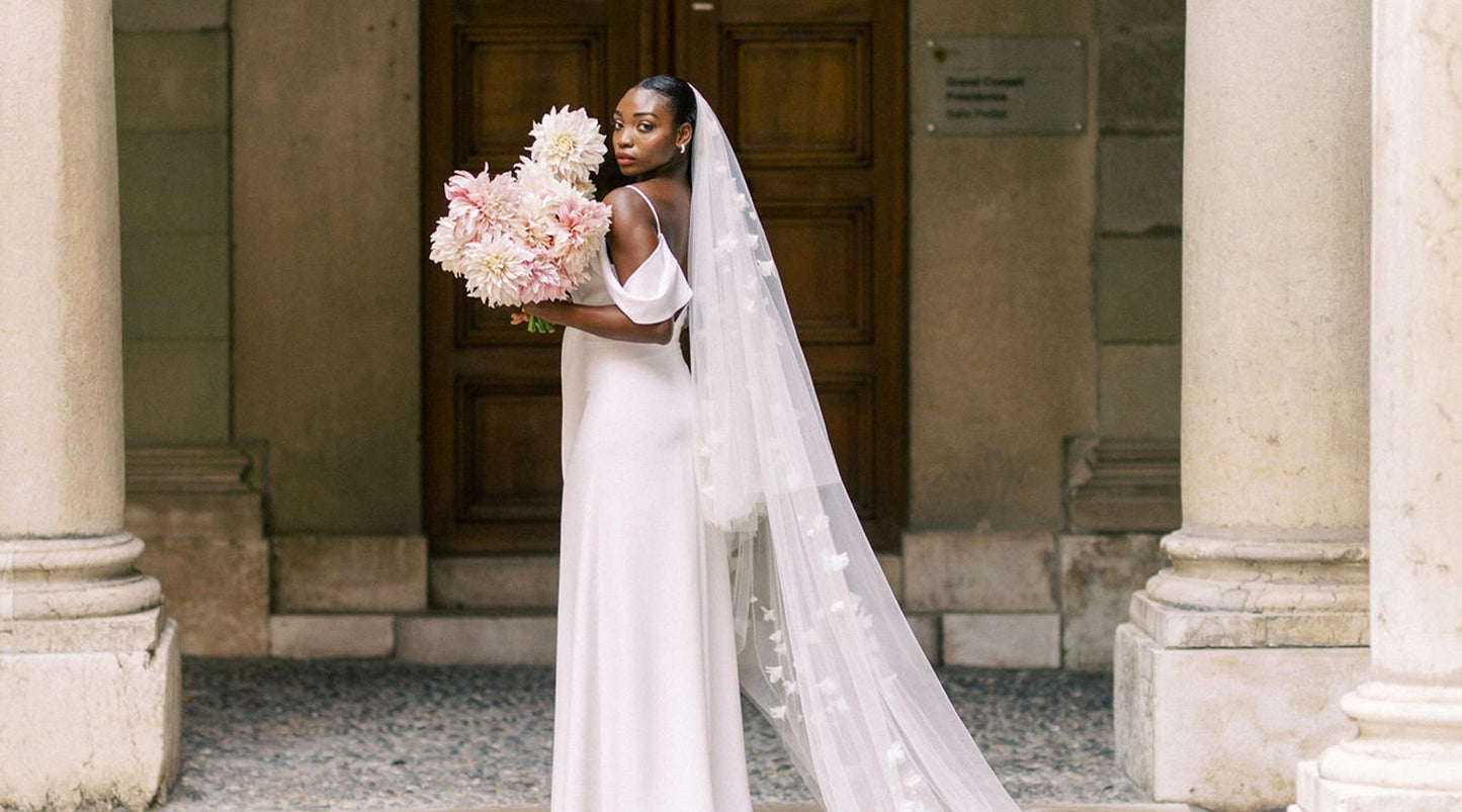 A bride wearing a plain white gown and a long floral wedding veil holding a bouquet of flowers