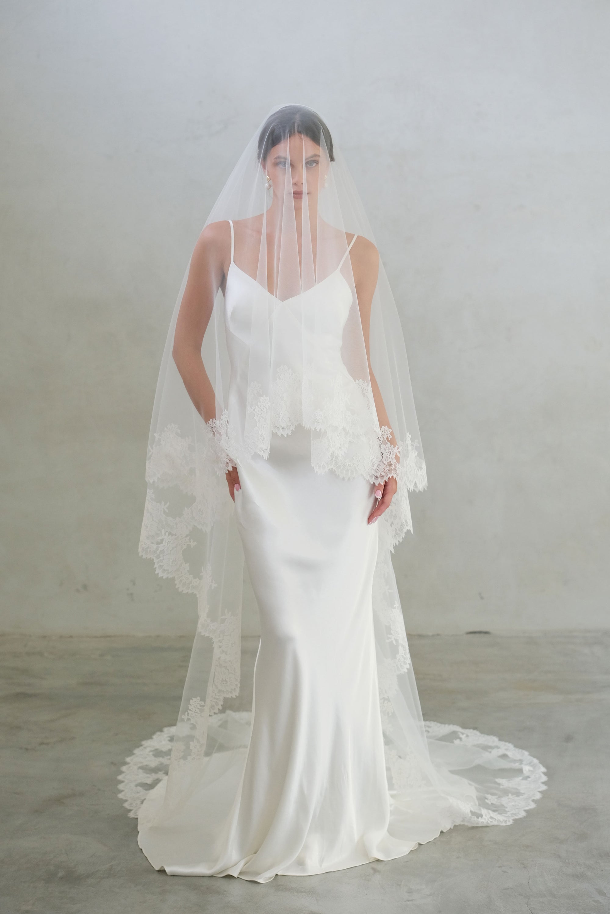 Full length front facing bride wears blusher down on two tier, chapel length wedding veil with elaborate lace trim