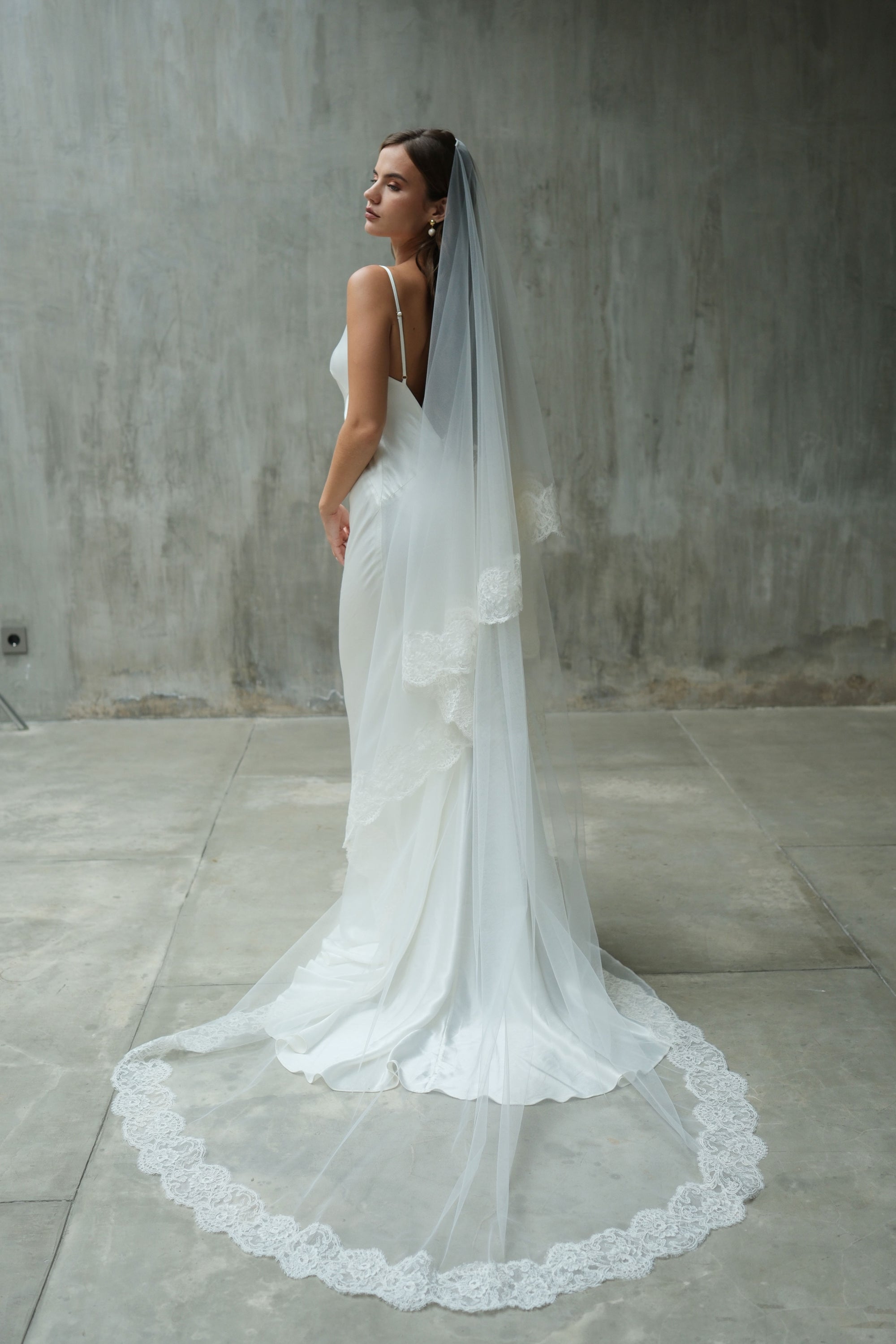 Full length bride in profile shows long veil with blusher off and train fanned out to display romantic lace edging