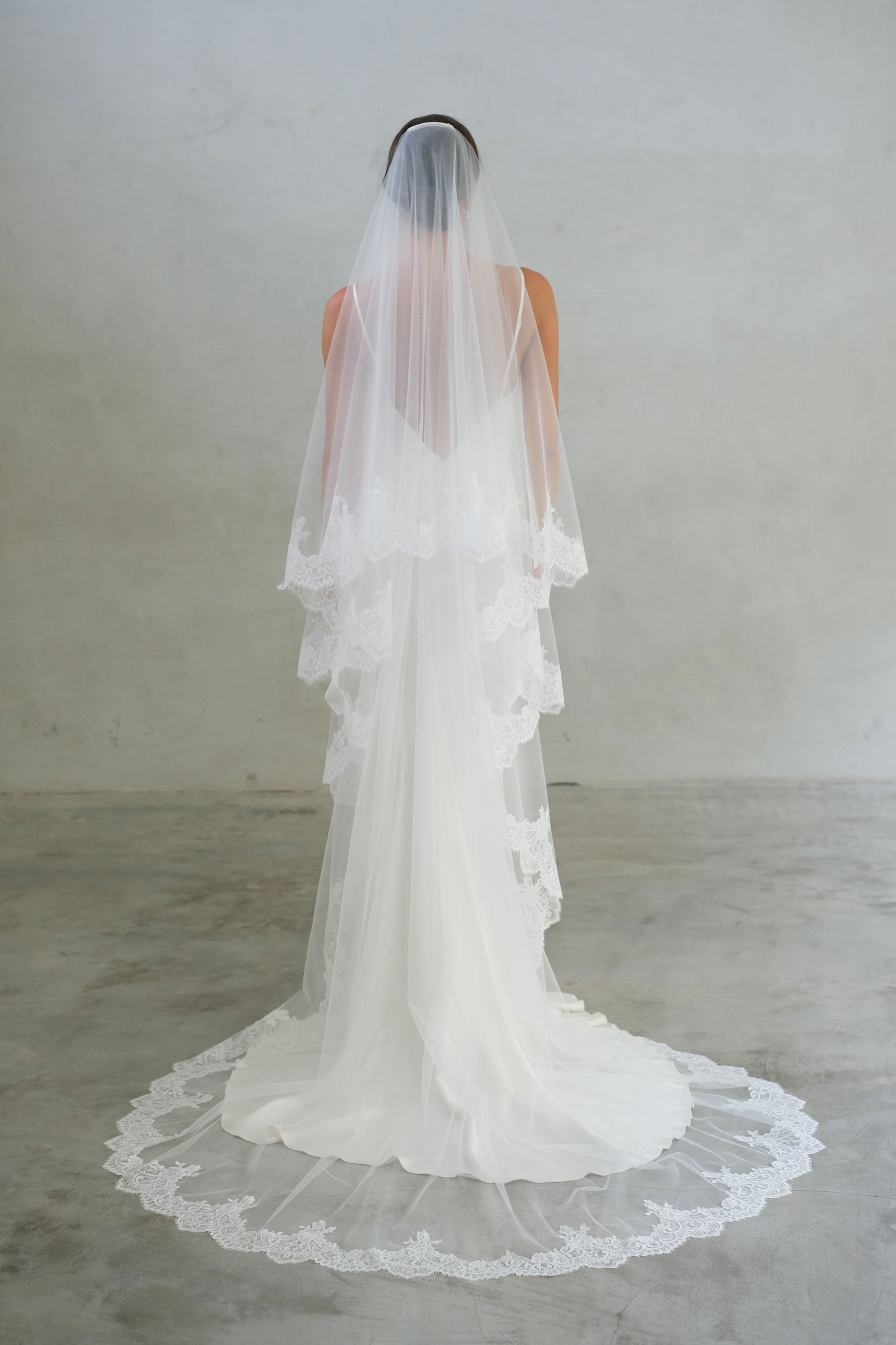 Full-length back view of a bride wearing two-tier lace trim long modern wedding veil in chapel length, blusher off