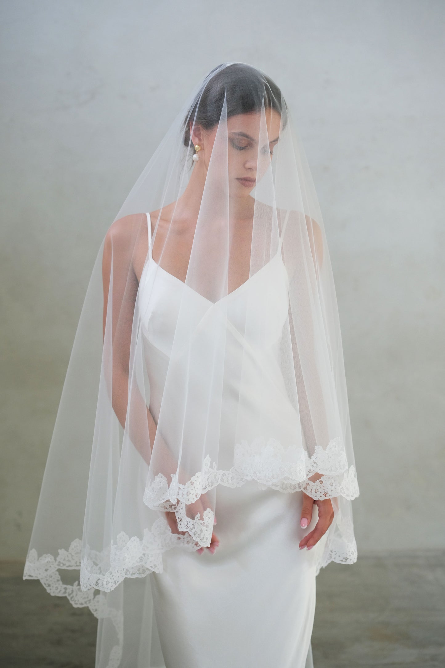 Half length front facing bride wears long laced edged drop veil, blusher down