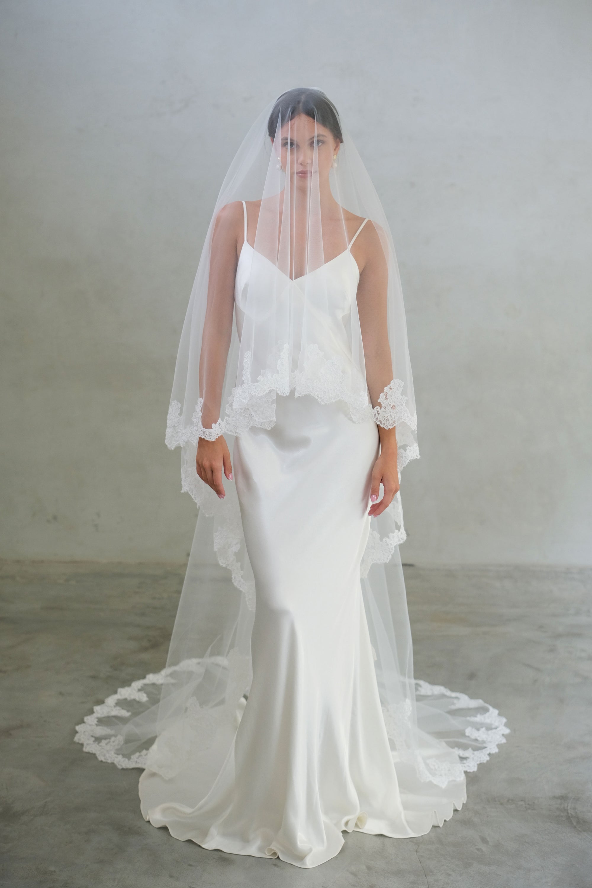 Full length front facing bride wears long, two-tier wedding veil with lace edging; blusher down