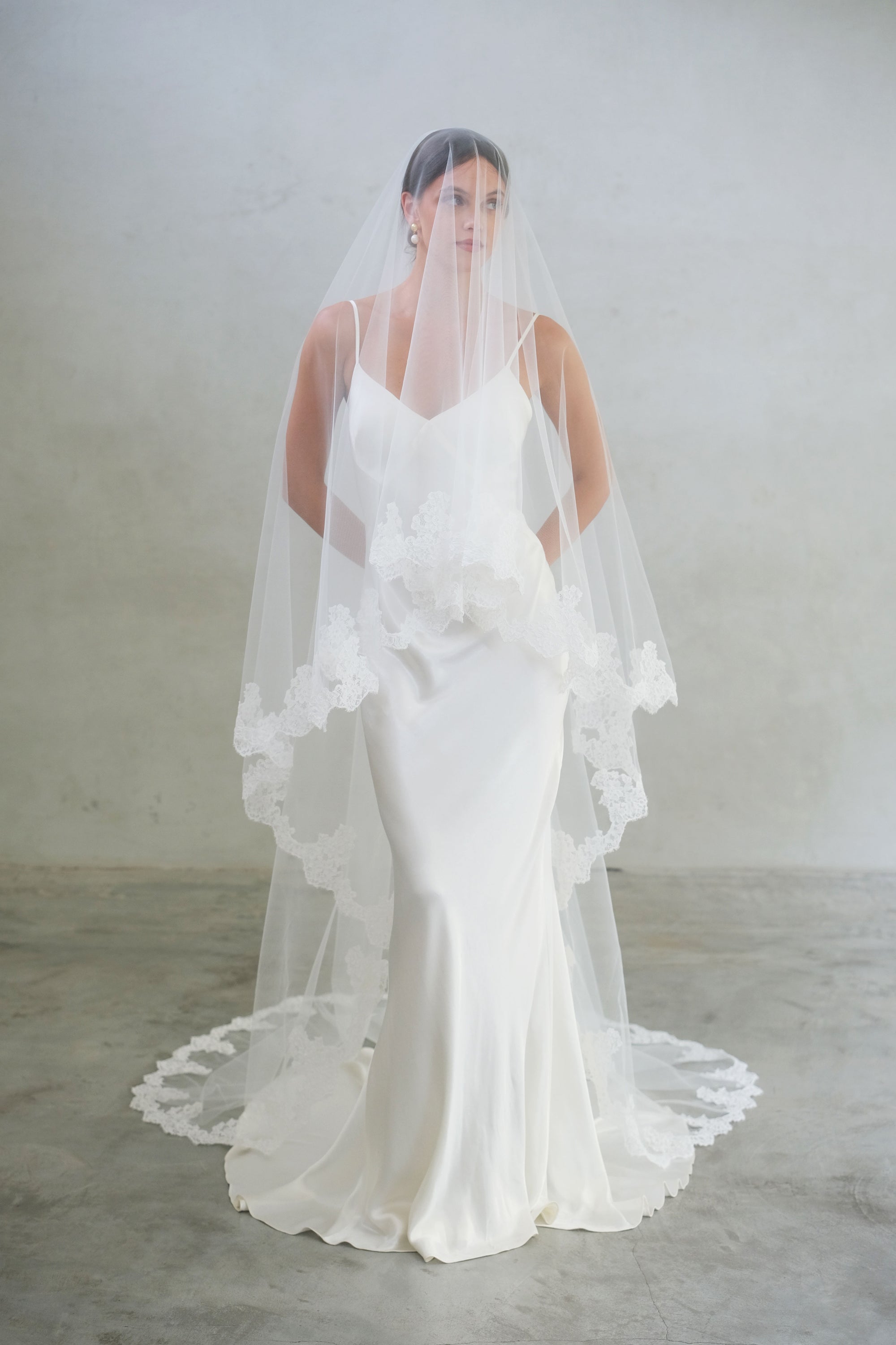 Full length front facing bride wears chapel length, lace trimmed wedding veil shown with blusher down