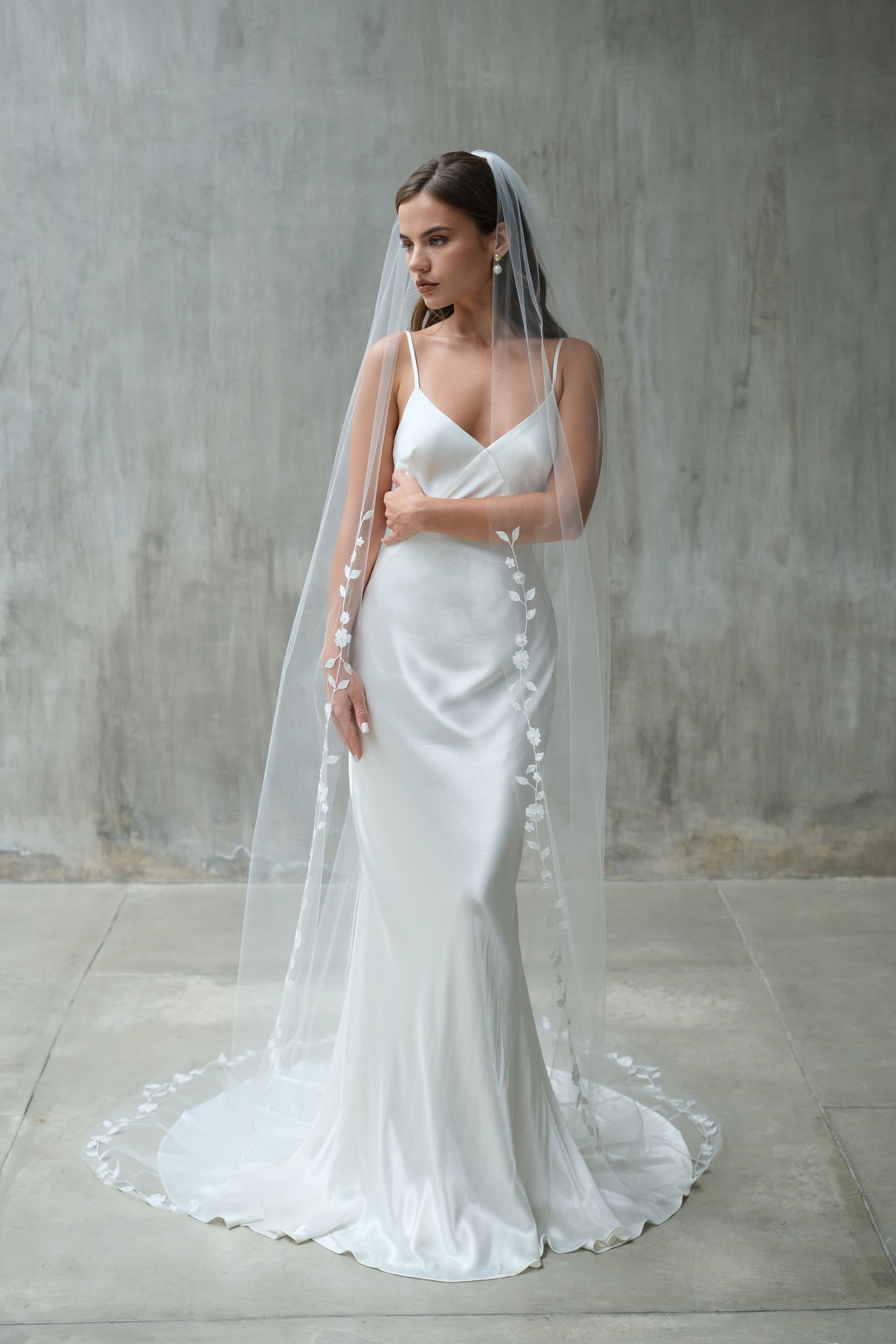 Full length front facing bride wears long veil, gathered at the comb and edged with delicate floral embroidery