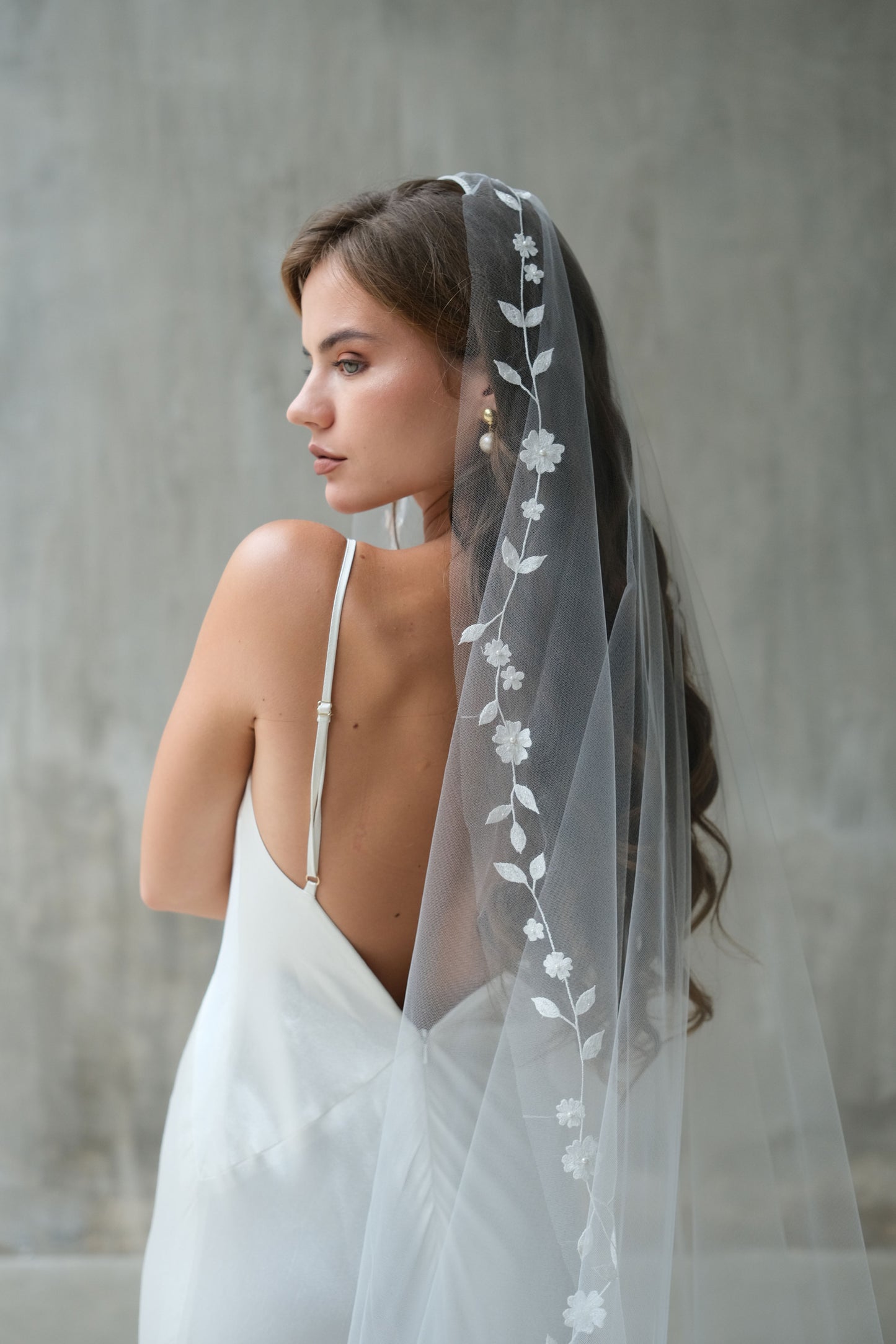 Bride in profile wears a long mantilla style wedding veil with floral embroidered edging and featuring minimal pearl beading