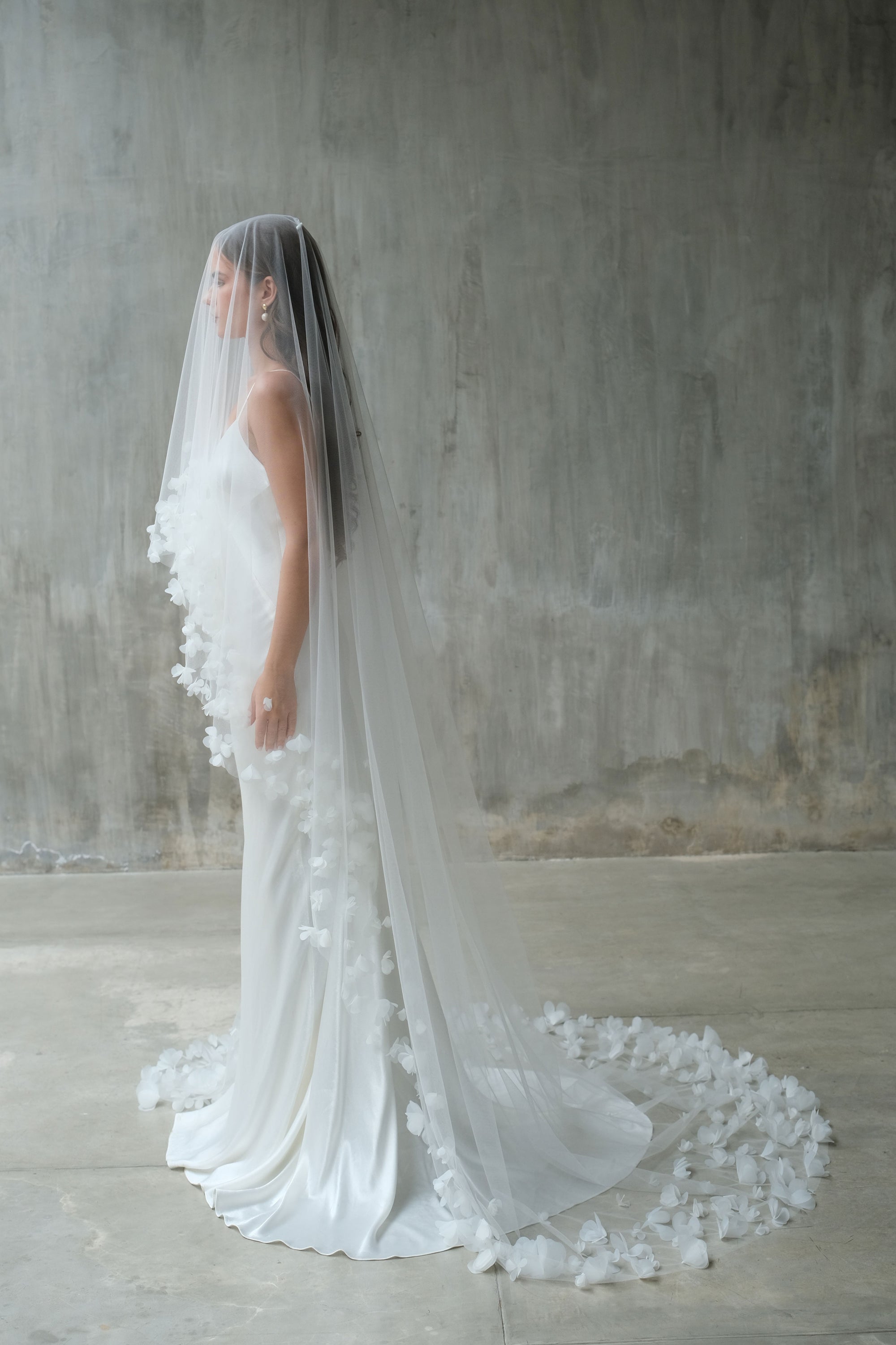 Full length side view of bride wearing long veil edged with fabric flowers, blusher down