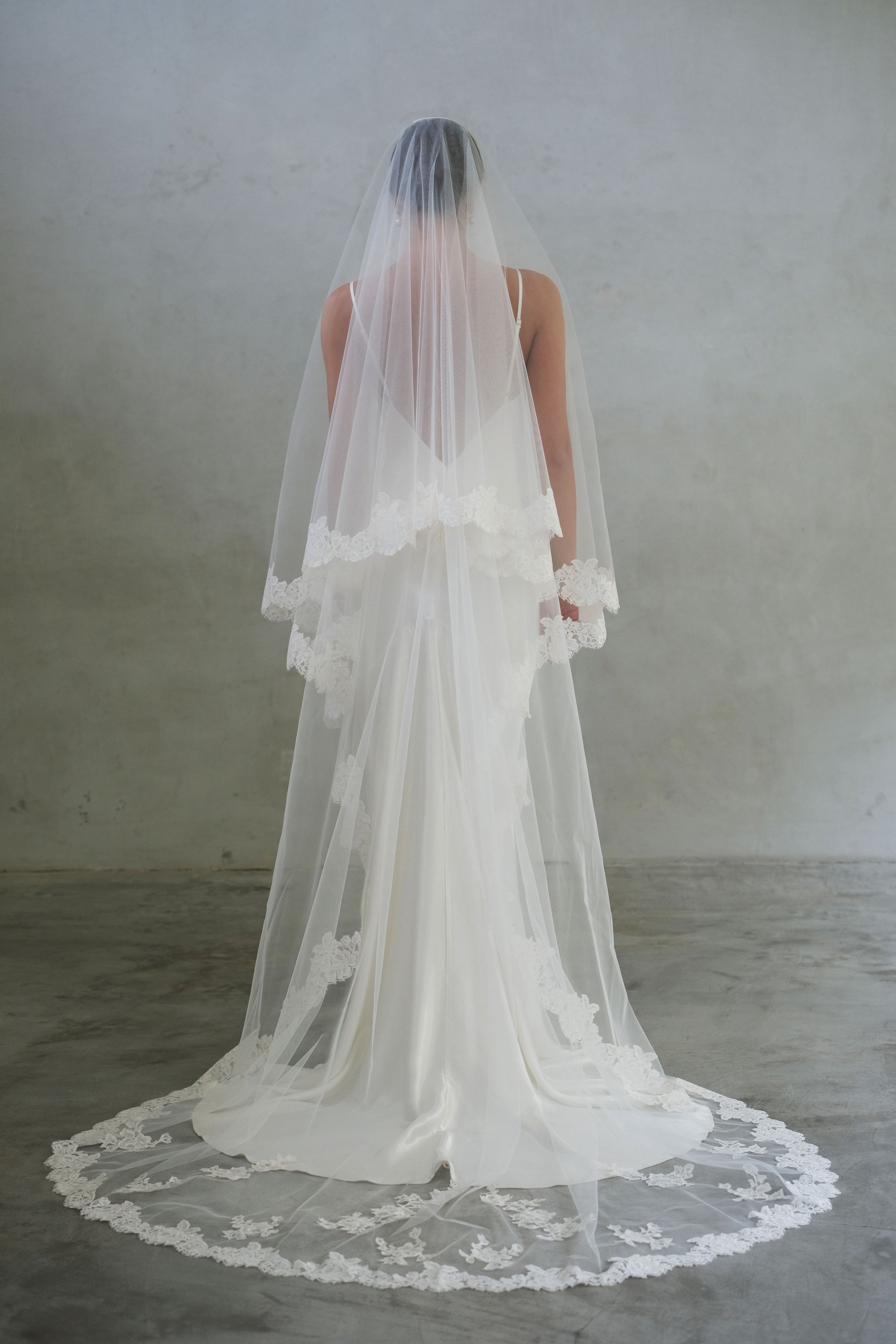Full-length back view of bride wearing long, lace-edged, two-tiered veil featuring scattered floral applique detail all around the train