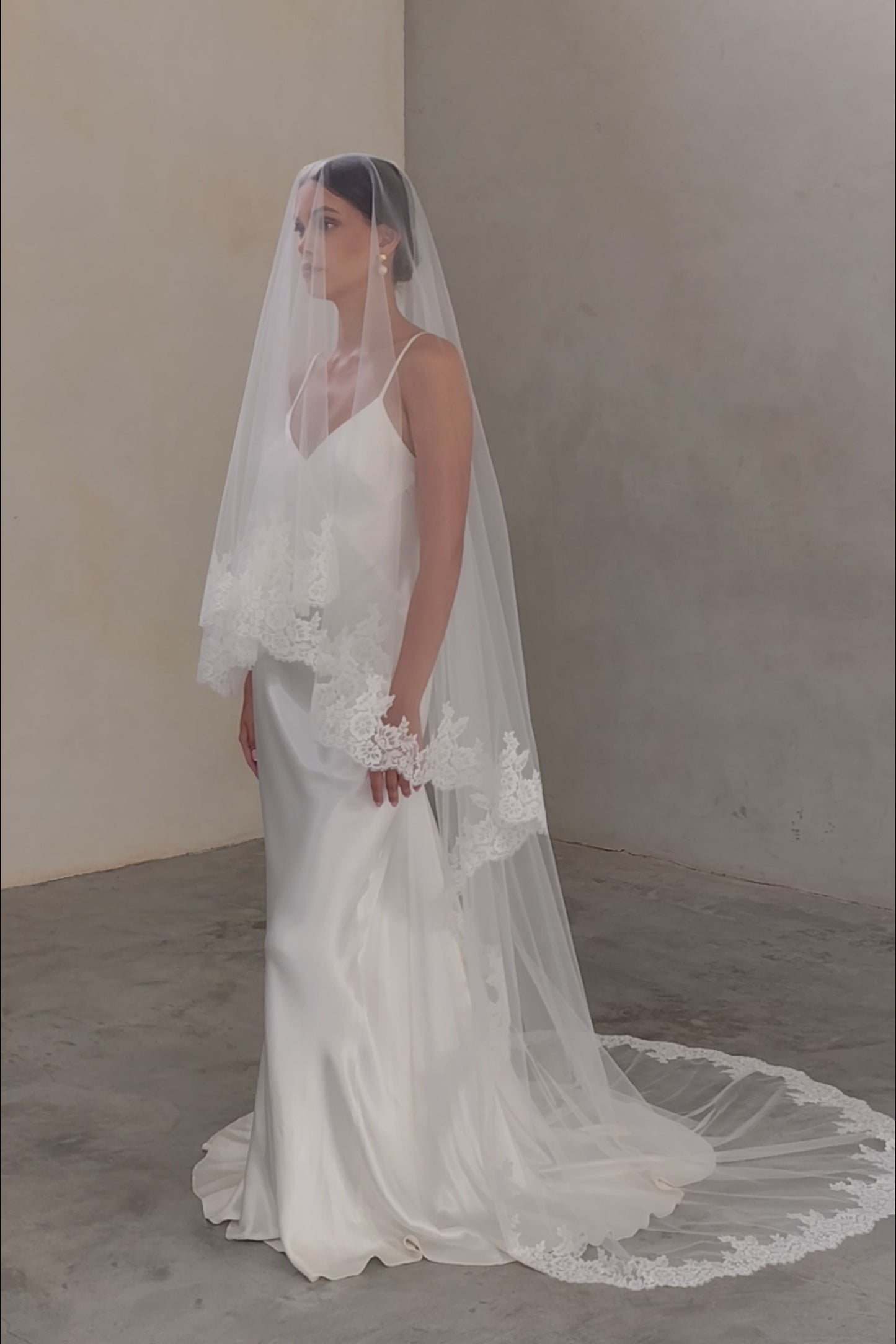 Full length side facing bride wears blusher down on two tier veil trimmed at the edge with romantic lace