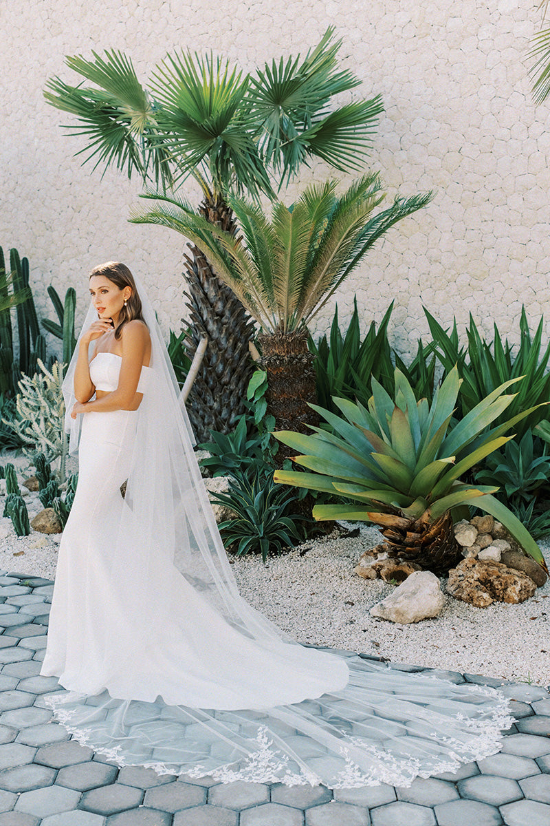 Full length side view of bride in palm garden setting wearing long, two tier wedding veil embroidered with floral design all around the train